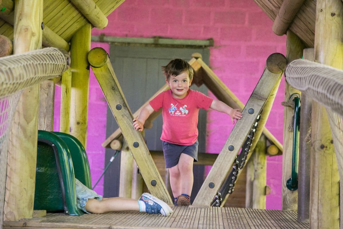 A young boy is climbing through a wooden fort. A second child is lying down in a green slide.