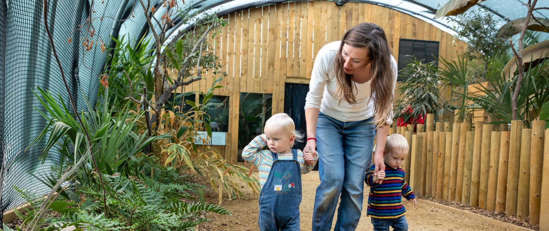 A woman walks hand in hand with two toddlers in a greenhouse-like setting, surrounded by plants and wooden fencing.