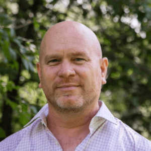 A headshot of a man in a purple shirt against a backdrop of greenery