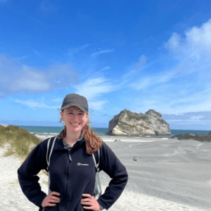 A person stood with hands on hips on a beach with a rocky outcrop behind them